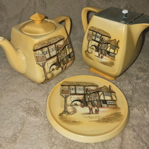 May include: A vintage tea set comprising a teapot, a creamer jug, and a small plate. The cream-coloured set features a detailed illustration of a building with the text "The Jolly Drover". The teapot has a lid and spout.