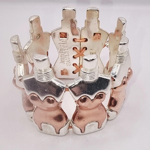Puede incluir: Pulsera plateada y oro rosa con un diseño repetido de frascos de perfume estilizados. La pulsera elástica presenta el texto "Jean Paul Gaultier". Fabricada en metal plateado y oro rosa.