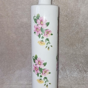 Può includere: Un mattarello in ceramica bianca caratterizzato da un motivo floreale con fiori rosa, foglie verdi e piccoli fiori gialli. Il corpo cilindrico presenta manici lisci e arrotondati su entrambe le estremità, con il disegno che prosegue sul manico.