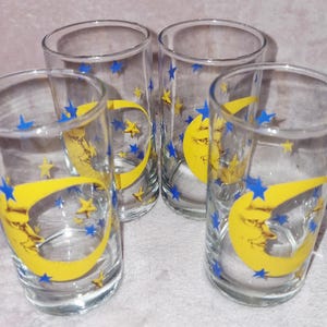 Puede incluir: Un juego de cuatro vasos de vidrio transparente, cada uno con un gráfico de luna creciente amarilla con cara y patrones de estrellas azules y amarillas dispersas impresos en la superficie exterior. Los vasos tienen forma cilíndrica.