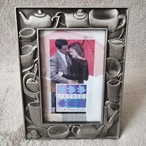 May include: A rectangular pewter-colored photo frame featuring a raised relief design of various teapots, cups, and saucers around the border. The frame holds a display card with the text SIXTREES and 4" x 6" Photo Frame, showing a couple.
