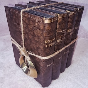 Könnte beinhalten: Ein Stapel aus vier Büchern in braunem Einband im Vintage-Stil, die mit natürlichem Jutegarn zusammengebunden sind. Ein polierter Messingring ist vorne am Garn befestigt. Die Buchrücken tragen goldene Schriftzüge mit Titeln wie Robinson Crusoe, The Three Musketeers, Westward Ho und Ivanhoe.