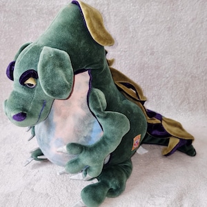 May include: A plush dragon toy featuring a dark green velvet body with purple accents on the eyes and nose. The creature has a light-colored, mottled belly, gold-toned fabric along the back and tail, and small, pointed silver-colored claws.