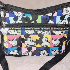 May include: A black crossbody bag featuring a colorful checkered pattern with repeating Mickey Mouse illustrations. The bag includes a front zippered pocket with a trim displaying the text Mickey Mouse in various colors and a silver Mickey-shaped zipper pull.