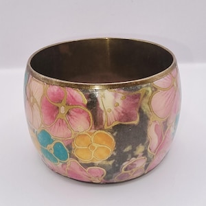 May include: A wide metal bangle bracelet featuring a colourful floral pattern with pink, yellow, and teal petals outlined in gold. The interior of the bracelet is a plain metallic finish, and the exterior surface is decorated with a repeating flower design.