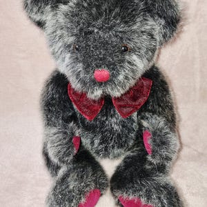 Könnte beinhalten: Ein Plüschteddybär mit schwarz-silbergrauem Fell, ausgestattet mit einer leuchtend roten Samtnase, einer passenden roten Samtfliege am Hals sowie roten Samtakzenten an den Fußsohlen und den Innenflächen der Vorderpfoten.