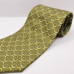 Pode incluir: Uma gravata com fundo verde-oliva e um padrão repetitivo de motivos em forma de ferradura em amarelo e dourado claro. O tecido tem uma textura de trama diagonal visível e a gravata está enrolada no topo para mostrar a estrutura interna.