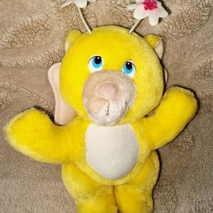Könnte beinhalten: Ein leuchtend gelbes Plüschtier mit beigefarbener Schnauze, blauen Augen und zwei Antennen, die mit weißen und rosafarbenen Stoffblumen verziert sind. Das Stofftier hat einen runden Bauchfleck, kleine Ohren und weiches Fell mit freundlichem Gesicht.