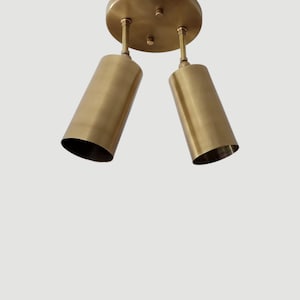 May include: A brass-colored, two-light ceiling fixture. The fixture has a circular base with two cylindrical lights suspended below. The lights are angled downward. The fixture is made of metal and has a warm, metallic finish.