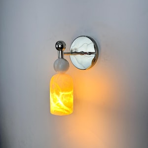 Può includere: Applique da parete con finitura cromata e luce giallo-arancio. Presenta una piastra posteriore rotonda, una piccola sfera, una sfera più grande e un paralume cilindrico con un motivo marmorizzato. La luce emana un bagliore caldo.