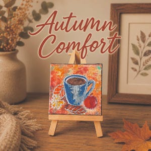 May include: A small square canvas painting on a wooden easel featuring a blue mug filled with a dark beverage, a red apple, and stylized autumn leaves in shades of orange and yellow. The text Autumn Comfort is written above the painting.
