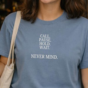 May include: A light blue t-shirt with the text "CALL. PAUSE. HOLD. WAIT. NEVER MIND." in white. The shirt is a crew neck style and is being worn by a person. A white tote bag is over the person's shoulder.