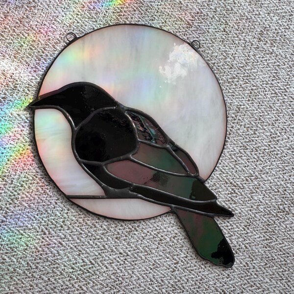 May include: Stained glass raven perched in front of a pearlescent moon. The raven is black with touches of green and burgundy. The moon is white with a subtle iridescent sheen. A small metal loop is at the top for hanging.