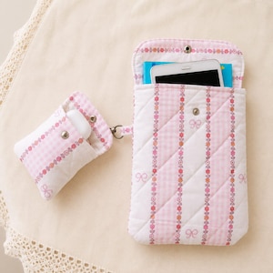 May include: A quilted fabric pouch set featuring a pink and white gingham pattern with vertical floral stripes and small bow motifs. The set includes a larger rectangular case with a snap closure containing a smartphone and a smaller matching case.