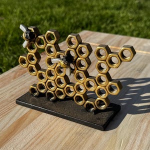 May include: A metal sculpture of a honeycomb with two bees. The honeycomb is made of gold-colored nuts and bolts, and the bees have black bodies and silver wings. The sculpture sits on a black rectangular base.