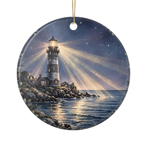 May include: A round ceramic ornament featuring a nighttime coastal scene with a lighthouse on a rocky shore. The lighthouse emits bright beams of light across a dark, starry sky and the ocean surface. A gold string is attached at the top.