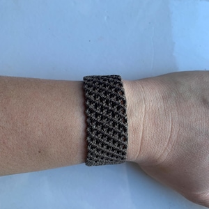 May include: A brown macrame bracelet is worn on a wrist. The bracelet is made of woven threads, creating a textured pattern. The bracelet is a wide band, and the color is a dark, earthy brown.