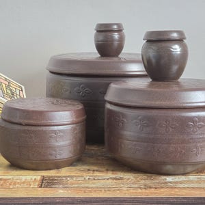 May include: Three dark brown ceramic storage jars with matching lids, each featuring a repeating embossed floral pattern around the sides. A small, square, multi-colored stained glass lamp sits to the left of the round, lidded pottery containers.
