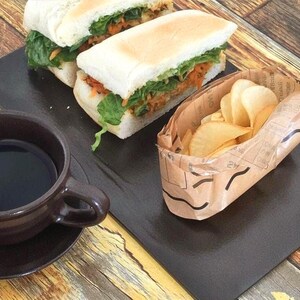 Könnte beinhalten: Zwei Weißbrot-Sandwiches gefüllt mit grünem Salat und geraspelten orangefarbenen Karotten, ein kleiner Papierbehälter mit dünnen Kartoffelchips und eine dunkelbraune Keramiktasse mit schwarzem Kaffee, alles auf einem dunklen Tablett.