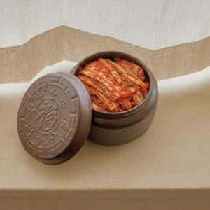 Könnte beinhalten: Ein dunkelbrauner Keramiktopf gefüllt mit leuchtend rotem Kimchi, begleitet von einem passenden runden Deckel mit einem geprägten Schriftzeichen in der Mitte und dekorativen Randmustern. Der Behälter hat eine matte Oberfläche und eine zylindrische Form.