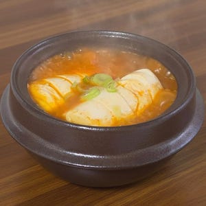 May include: A steaming bowl of Korean kimchi jjigae, a spicy stew with tofu and scallions. The stew is a vibrant orange color, and the tofu is a creamy white. The bowl is dark brown and sits on a wooden table.