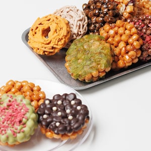 May include: An assortment of colorful, glazed pastries on a metal tray and a clear plate. The pastries vary in shape and color, including orange, brown, green, and chocolate. Some are topped with sprinkles or decorative elements.