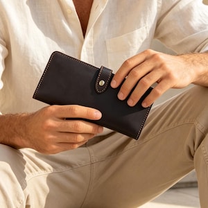 May include: A dark brown leather wallet featuring a rectangular shape, visible contrast stitching along the edges, and a secure flap closure with a small metallic snap button fastener centred on the front of the accessory.