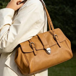 May include: A tan leather satchel bag with a rectangular shape and a flap closure. The bag features silver-coloured buckles and a shoulder strap. The bag is carried over the shoulder.