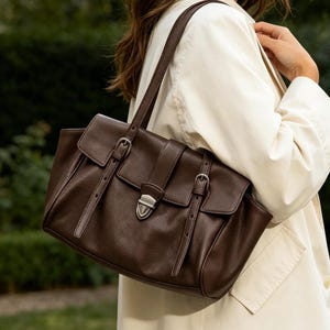 May include: A dark brown leather handbag with a rectangular shape. The bag features two front flaps secured with silver buckles and straps. The bag has a top handle and a shoulder strap. The background is a blurred green outdoor scene.