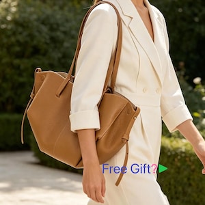 May include: A tan leather tote bag with long straps is carried over the shoulder. The bag has a unique, angular design and is made of a soft, supple leather. The image also shows a smaller matching bag. The text "Free Gift?" is visible in the image.