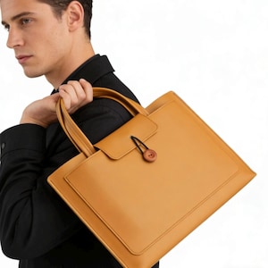 May include: A tan leather tote bag with a rectangular shape and a small flap closure secured by a wooden button. The bag has two handles and a pocket on the front. The bag is carried over the shoulder.