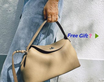 Op de afbeelding: Een beige handtas met een getextureerd oppervlak, een enkel handvat en een verstelbare schouderriem. De tas heeft een klepsluiting met een zilverkleurig metalen draaislot. De tekst "Free Gift?" is zichtbaar in het blauw en roze.