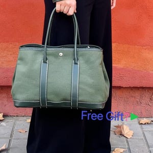 Pode incluir: Uma bolsa tote de lona verde-oliva com acabamento em couro verde-escuro, duas alças superiores e um fecho central de botão de pressão prateado. A bolsa apresenta costuras brancas visíveis nos detalhes de couro e o texto Free Gift com uma seta verde.