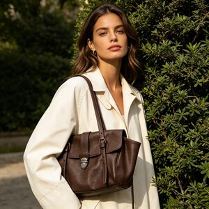May include: A dark brown leather satchel bag with a flap closure and silver-tone hardware. The bag has a top handle and a shoulder strap. The bag is worn over the shoulder. The background is a green bush and a person wearing a white coat.
