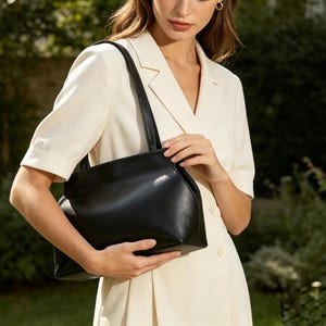 May include: A black leather tote bag with a rectangular shape and a shoulder strap. The bag is held by a person wearing a cream-colored dress. The background features green foliage.