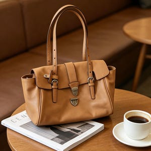 May include: A tan leather handbag with a rectangular shape, featuring a flap closure secured by a metal clasp and two buckles. The bag has two top handles and is sitting on a wooden table next to a cup of coffee and a magazine.