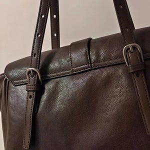 May include: Close-up of a brown leather handbag with adjustable straps and silver-coloured buckles. The bag has a flap closure with a buckle and a top handle. The leather appears smooth and has a slight sheen.