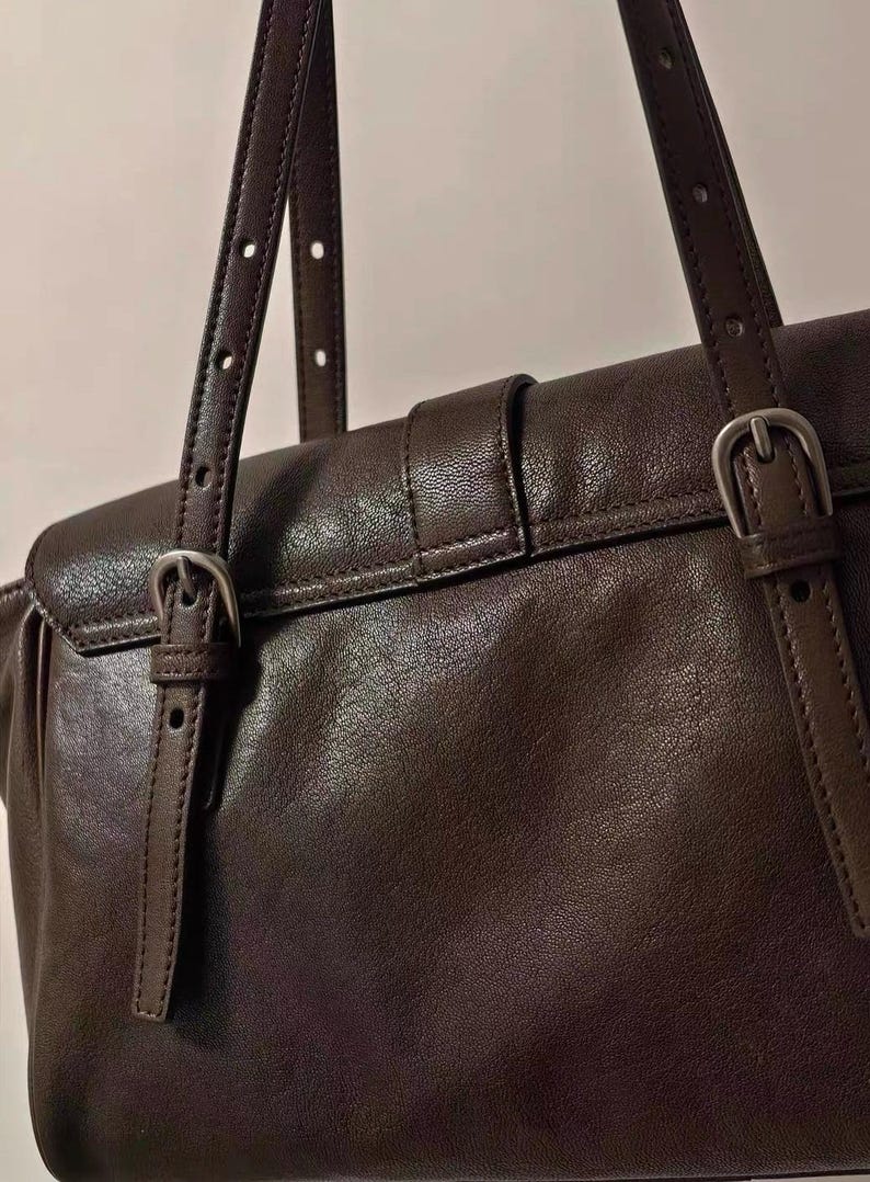 May include: Close-up of a brown leather handbag with adjustable straps and silver-coloured buckles. The bag has a flap closure with a buckle and a top handle. The leather appears smooth and has a slight sheen.