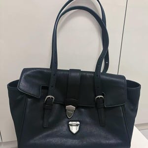May include: A black leather tote bag with two top handles and a flap closure. The bag features silver-coloured buckles and hardware. The design includes a front flap with a buckle closure and two side pockets.