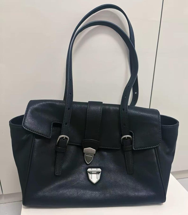May include: A black leather tote bag with two top handles and a flap closure. The bag features silver-coloured buckles and hardware. The design includes a front flap with a buckle closure and two side pockets.