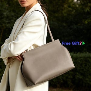 May include: A taupe-colored handbag with a rectangular shape and a long, thin strap. The bag has a curved design on the sides and is held by a person wearing a white blazer. The text "Free Gift?" is visible.