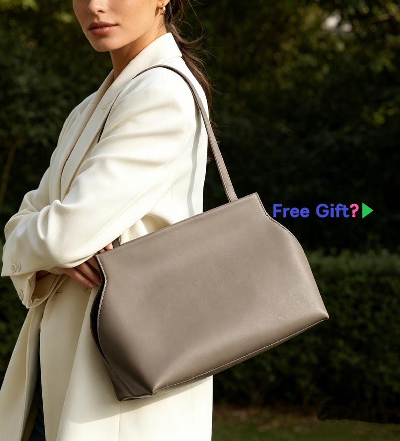 May include: A taupe-colored handbag with a rectangular shape and a long, thin strap. The bag has a curved design on the sides and is held by a person wearing a white blazer. The text "Free Gift?" is visible.