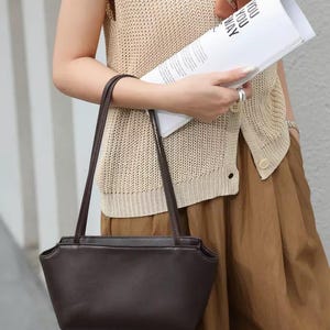 以下が含まれることがあります： 長方形の底と先細りの上部を持つダークブラウンのレザーショルダーバッグ。2本の細い同色のブラウンのショルダーストラップと、滑らかでマットな質感が特徴です。シンプルで構造的なデザインで、すっきりとしたシルエットをしています。