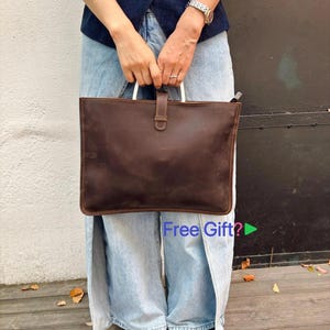 Könnte beinhalten: Eine braune Ledertasche mit weißen Griffen und einem Schnallenverschluss. Die Tasche ist rechteckig und wird von einer Person getragen. Die Tasche hat ein schlichtes, klassisches Design.