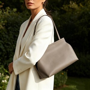 May include: A gray leather handbag with a rectangular shape and a single shoulder strap. The bag is carried by a person wearing a white blazer. The background is a blurred green foliage.