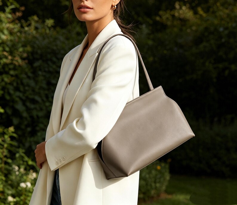 May include: A gray leather handbag with a rectangular shape and a single shoulder strap. The bag is carried by a person wearing a white blazer. The background is a blurred green foliage.