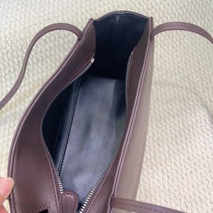 以下が含まれることがあります： 細いストラップとシルバーカラーの金属製ジッパーが付いたダークブラウンのショルダーバッグ。バッグの内側は滑らかな黒い素材で裏打ちされており、外側は同色のステッチが施された無地のダークブラウン仕上げになっています。