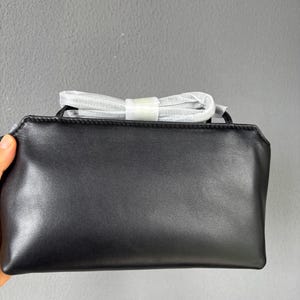 以下が含まれることがあります： 滑らかな仕上げの長方形の黒いレザー製クラッチバッグで、上部にファスナーが付いています。バッグには、白い保護紙で包まれた細い折り畳み式のショルダーストラップが付いており、メインコンパートメントの両サイドに固定されています。