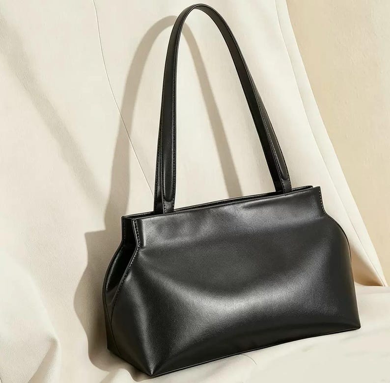 May include: A black handbag with two long handles. The bag has a rectangular shape with rounded corners and a smooth, shiny surface. The handles are thin and attached to the top of the bag. The background is a light color.