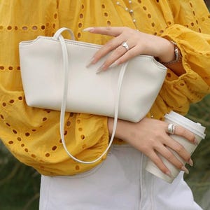 以下が含まれることがあります： 細いストラップが付いた滑らかなオフホワイトの長方形のショルダーバッグ。白い使い捨てコーヒーカップと一緒に持たれています。バッグは、上部にジッパー開閉部と湾曲した上端のデザインを備えた、すっきりとしたシルエットが特徴です。